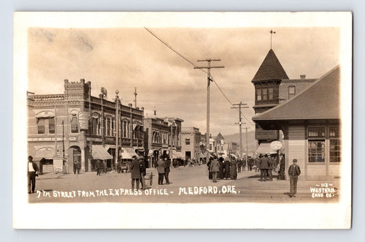 Sepia real photo postcard showing 7th Street in downtown Medford, Oregon, with storefronts, pedestrians, and towered building at right; unposted divided back.