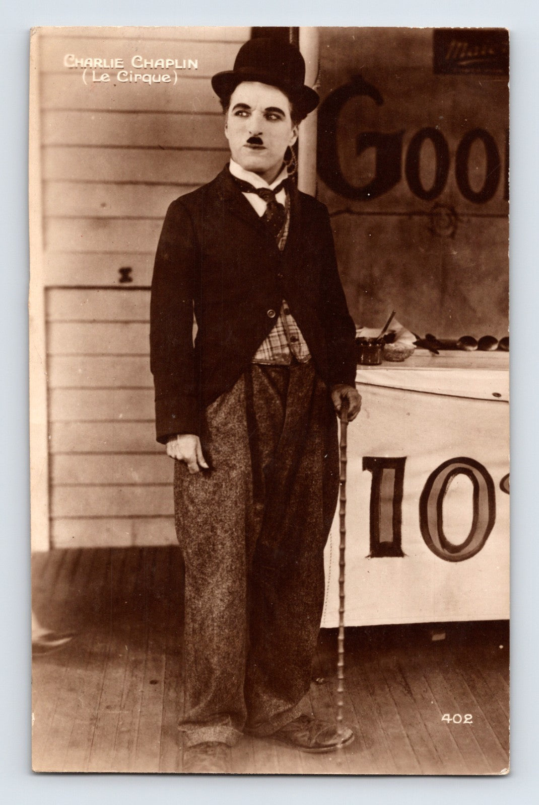 Sepia real photo postcard showing Charlie Chaplin in bowler hat and cane from Le Cirque standing beside fairground booth backdrop; unposted real photo back.