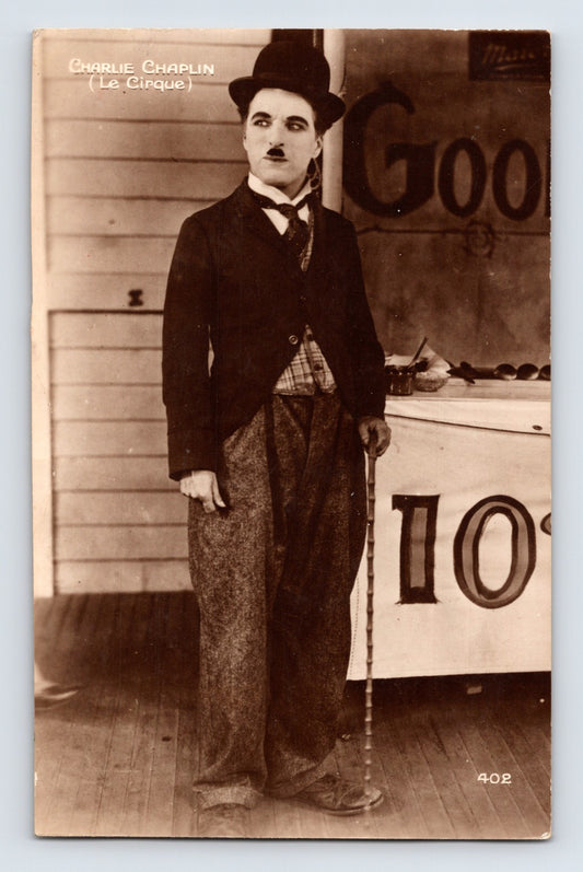 Sepia real photo postcard showing Charlie Chaplin in bowler hat and cane from Le Cirque standing beside fairground booth backdrop; unposted real photo back.