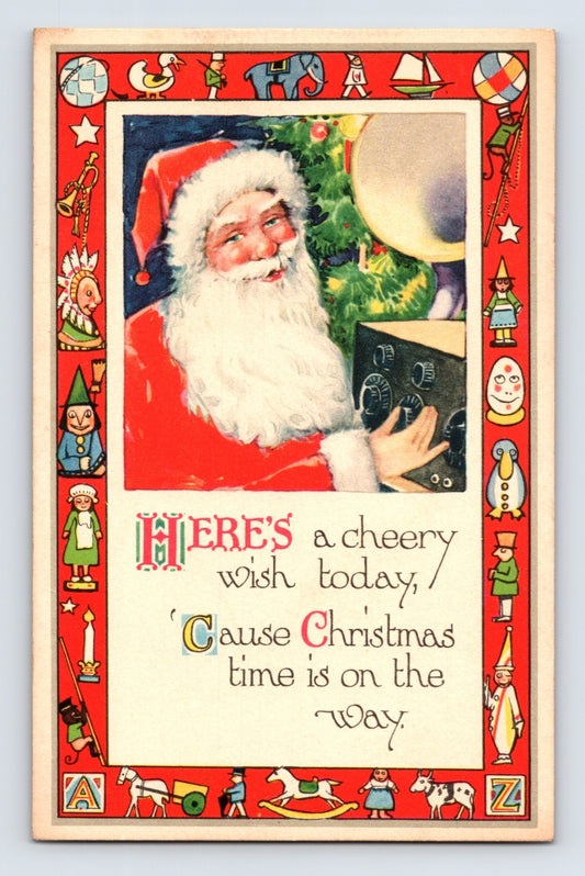 Santa Claus beside a Christmas tree adjusting a large radio or phonograph console, framed by a bright red border filled with toys and nursery figures.