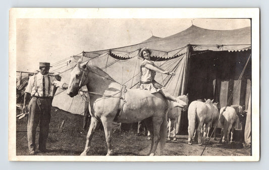Black and white photograph showing a female circus performer in costume seated on a white horse beside a tent with additional horses and handler visible