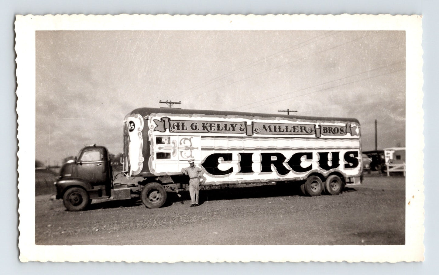 Black and white photograph of Kelly Miller Bros Circus truck and trailer with large painted lettering and driver standing beside vehicle on open lot