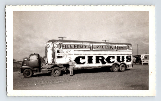 Black and white photograph of Kelly Miller Bros Circus truck and trailer with large painted lettering and driver standing beside vehicle on open lot