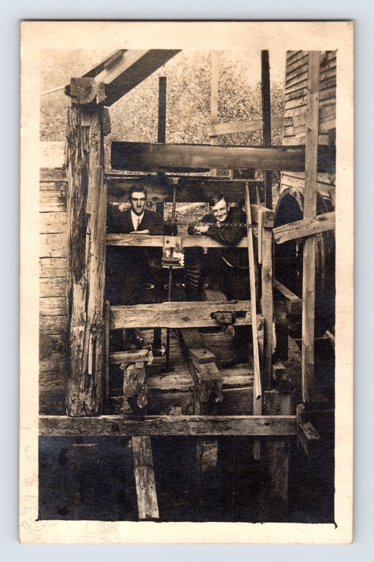 1910s real photo postcard of interior mill machinery in Lowville, New York with exposed gears, belts, timber framing, and two men posed inside the structure