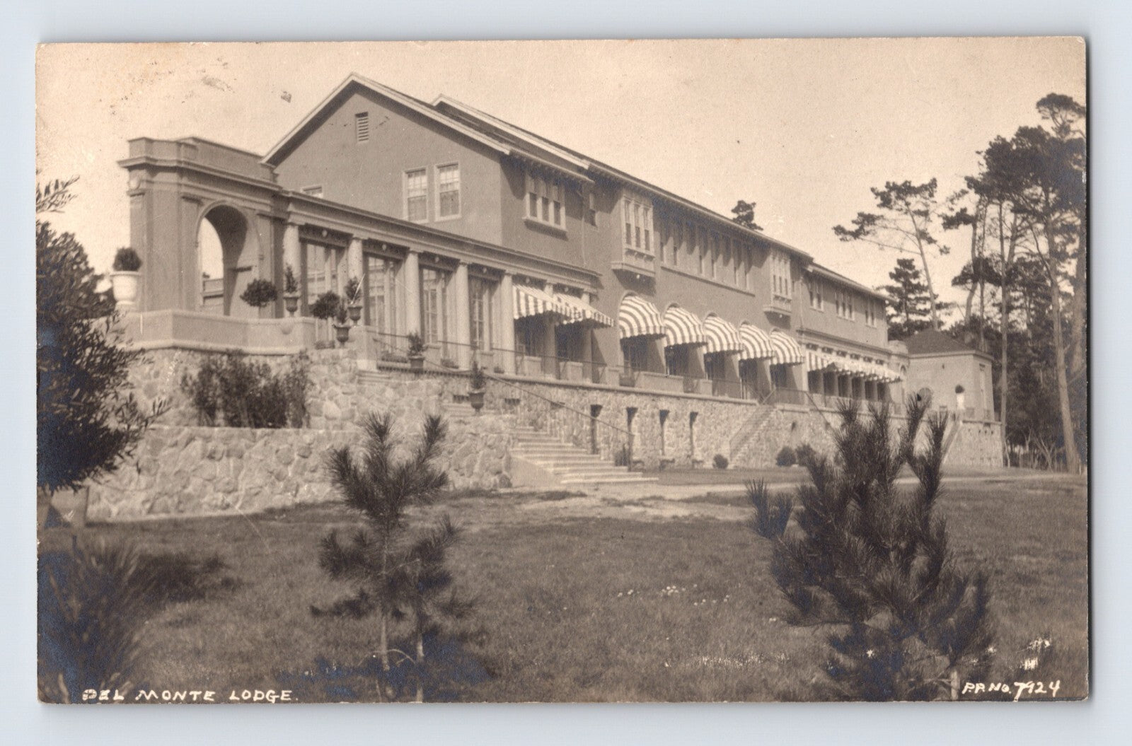 Sepia-tone real photo postcard of the Del Monte Lodge at Pebble Beach, California, showing the large resort building with terraces, striped awnings, and landscaped grounds.