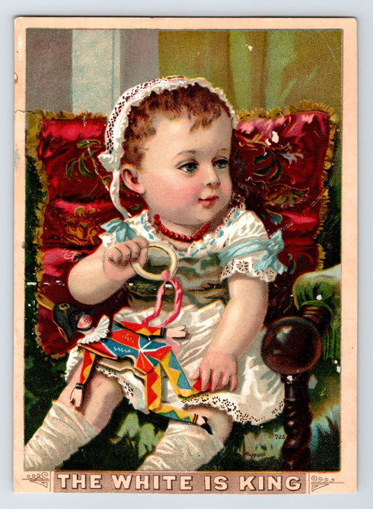 1890s Victorian trade card for White Sewing Machine showing baby seated with toy harlequin doll in richly colored interior scene