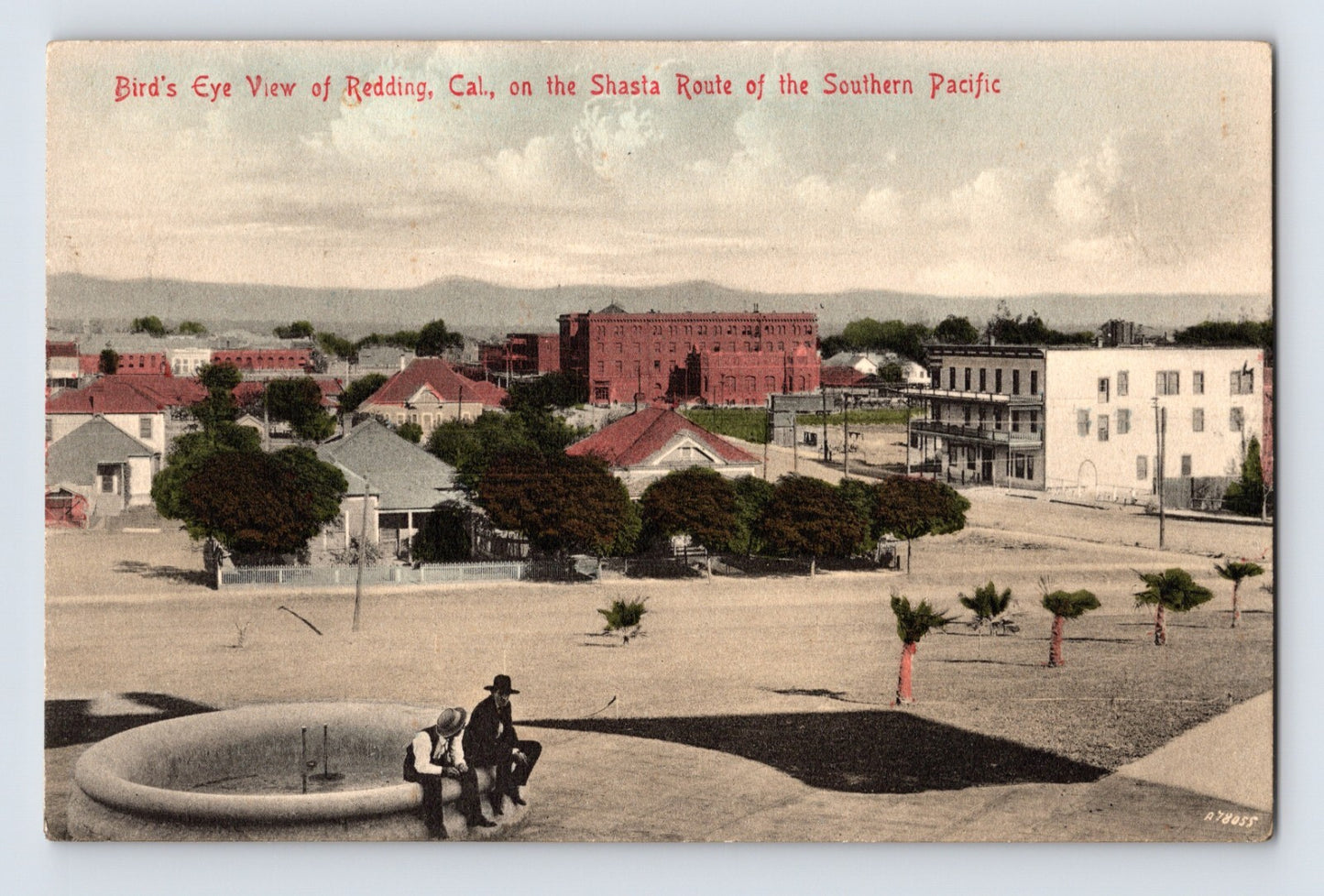 Hand colored early 1910s postcard showing bird’s eye view of downtown Redding, California with low buildings, red brick block, distant mountains, and fountain in foreground