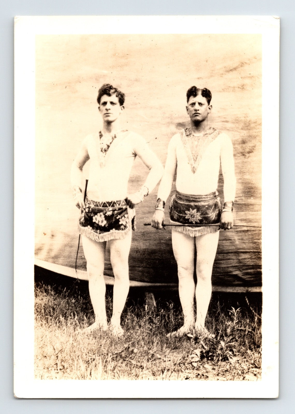 Sepia photograph of two circus performers in costume holding riding whips standing barefoot outdoors