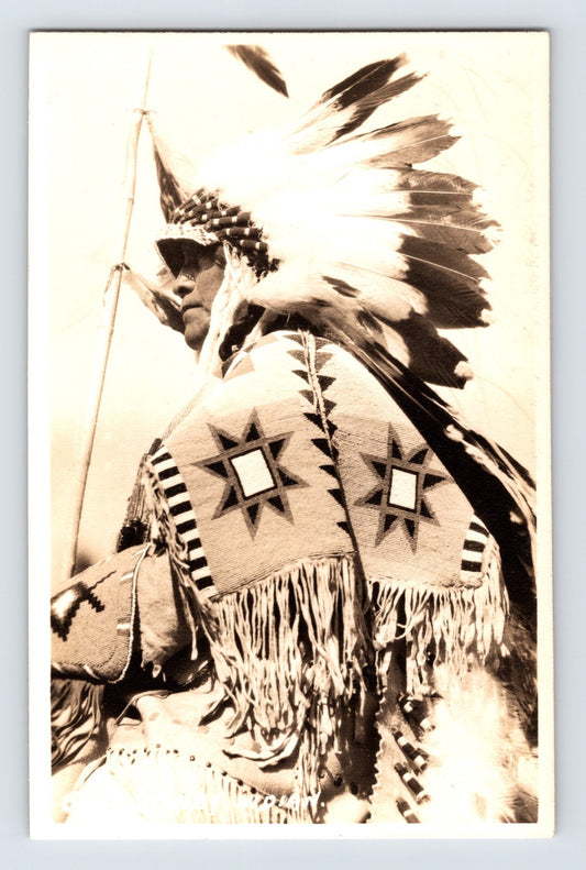 postcard front showing a Stoney Nakoda man in profile wearing a feathered war bonnet and fringed beaded garment with star motifs; Western Canada, unposted 1930s real photo.