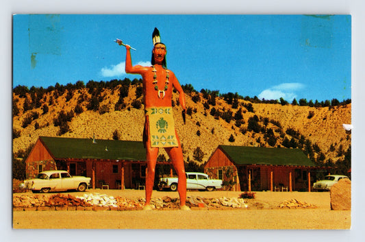 1960s postcard showing a giant Native American roadside statue at Fisher Rancho Lodge in Orderville, Utah, with motel cabins, vintage automobiles, and desert hills in the background.