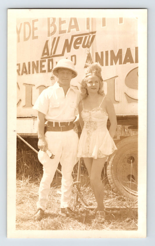 Sepia photograph showing a Clyde Beatty Circus animal trainer and female showgirl identified as Harriet standing before circus wagon signage