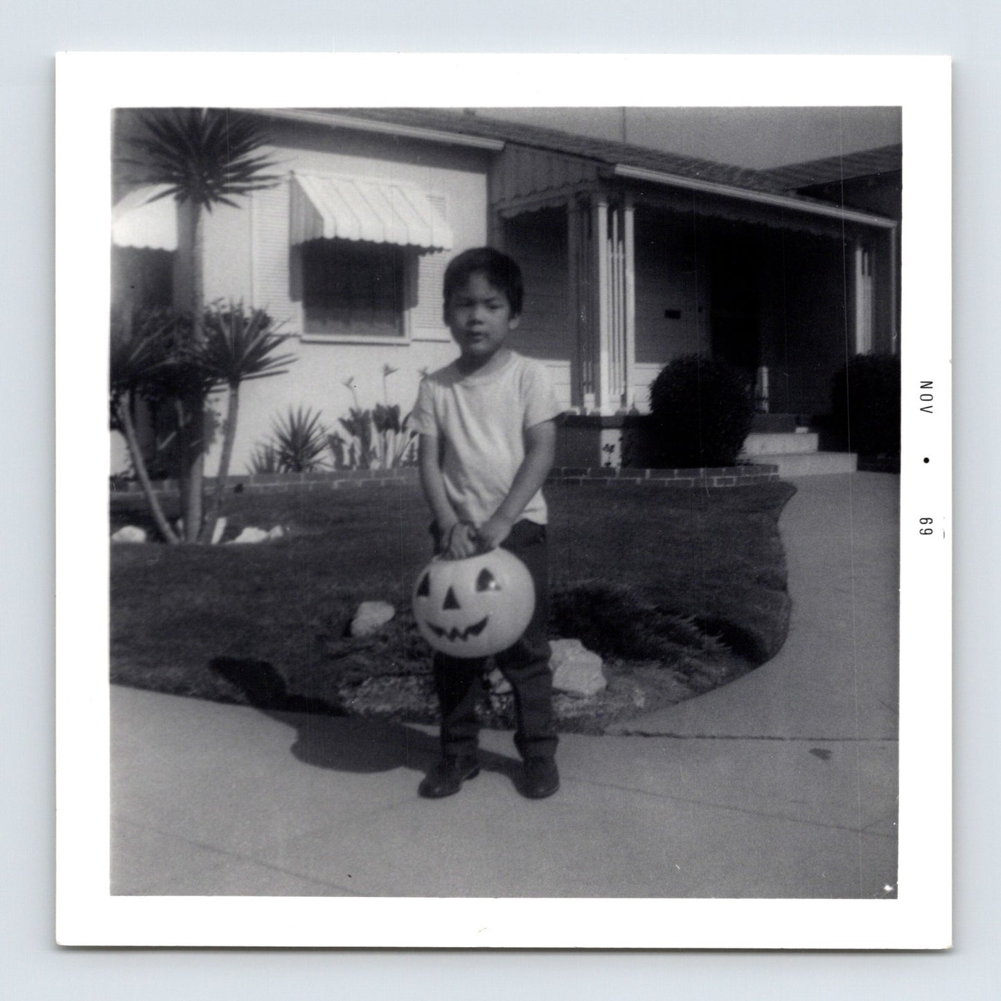 Halloween snapshot of young child holding jack o lantern trick or treat bucket in front of mid century suburban house daytime 3.5 x 3.5 photo