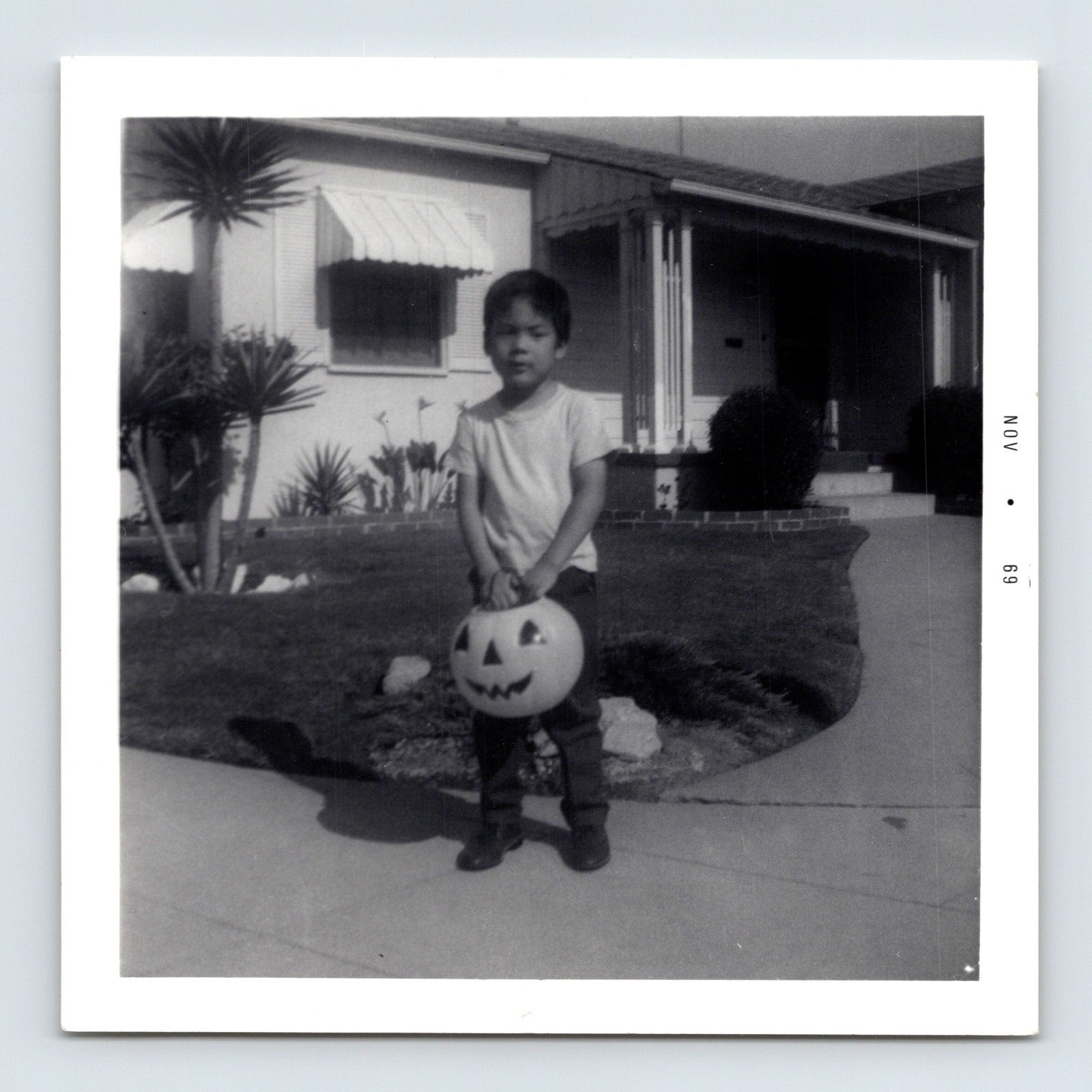Halloween snapshot of young child holding jack o lantern trick or treat bucket in front of mid century suburban house daytime 3.5 x 3.5 photo