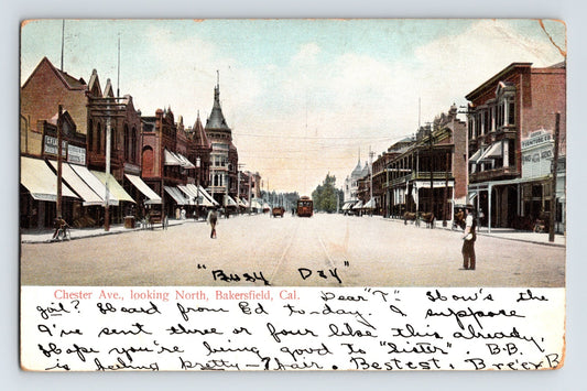 1906 Chester Avenue Bakersfield California postcard showing brick storefronts, awnings, early automobiles, pedestrians, and handwritten message across the lower margin.