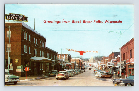 1962 downtown Black River Falls Wisconsin postcard showing Main Street storefronts, parked 1950s automobiles, Freeman Hotel sign, and Welcome Hunters banner spanning the street.
