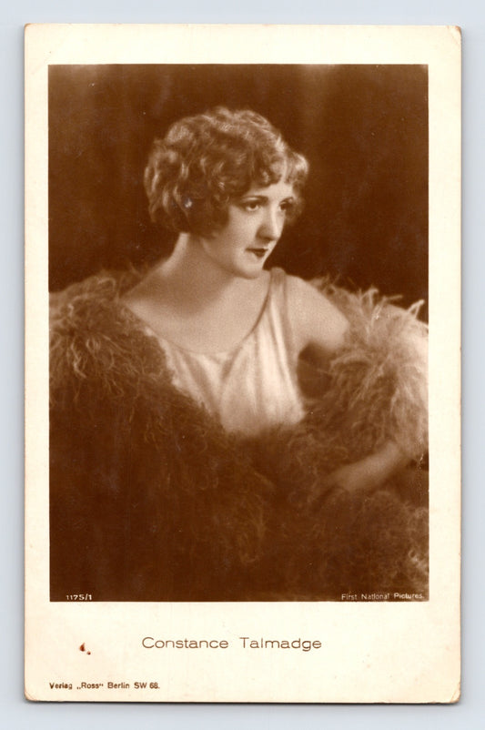 Sepia-tone portrait postcard of silent film actress Constance Talmadge wearing a satin dress and feathered wrap in a dramatic studio publicity photograph.