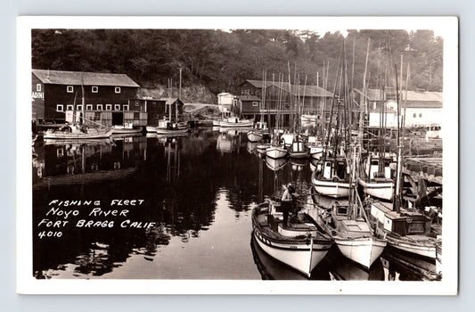 Black and white real photo postcard showing fishing boats moored in Noyo Harbor with waterfront buildings and wooded hillside behind, captioned Fishing Fleet Noyo River Fort Bragg Calif.