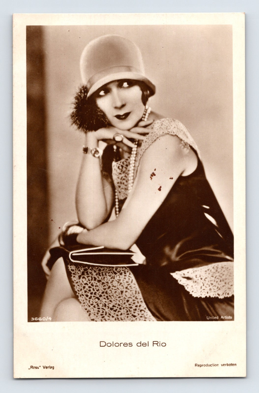 Sepia-tone portrait postcard of silent film actress Dolores del Rio wearing a cloche hat, pearls, and fashionable evening attire in an Art Deco studio pose.