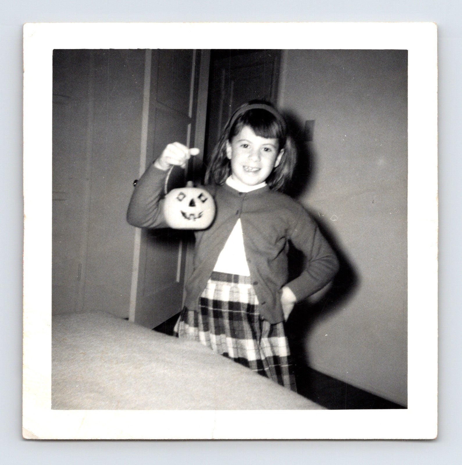 Girl holding jack o lantern pumpkin pail indoors wearing plaid skirt and cardigan Halloween snapshot 1960s