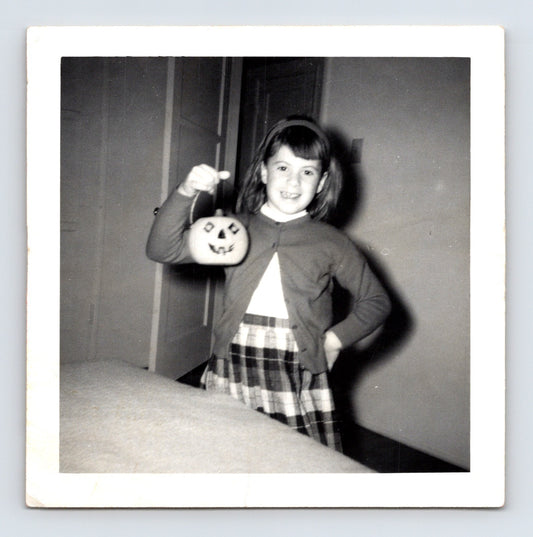Girl holding jack o lantern pumpkin pail indoors wearing plaid skirt and cardigan Halloween snapshot 1960s