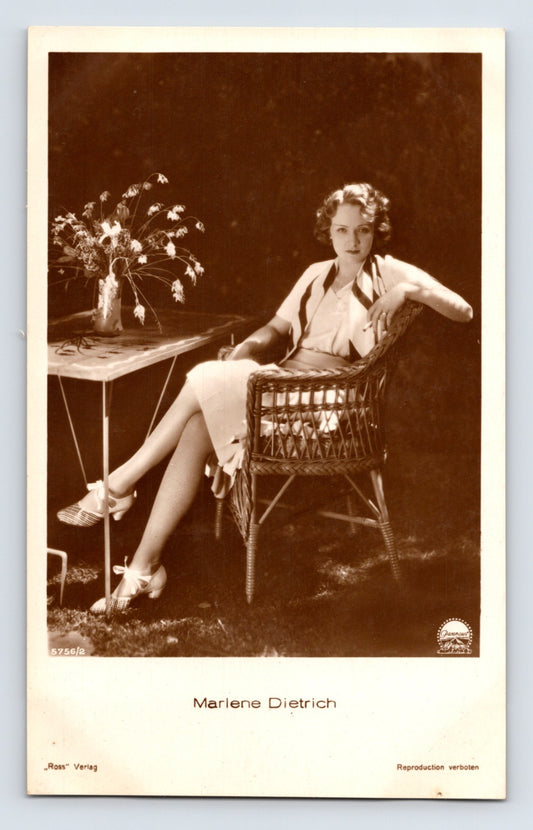Real photo postcard portrait of Marlene Dietrich seated in wicker chair beside small table, 1930s Ross Verlag studio image