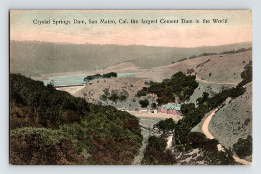 Hand-colored postcard showing Crystal Springs Dam in San Mateo County, California, with reservoir, rolling hills, roadway, and small buildings in foreground; divided back format.