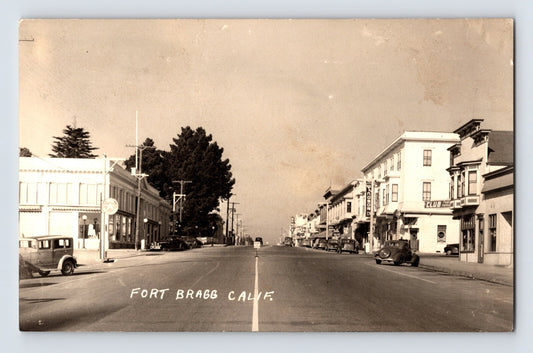 Black and white real photo postcard showing a wide street view of downtown Fort Bragg, California, with storefronts, parked automobiles, and signage including Windsor Cafe and Club.