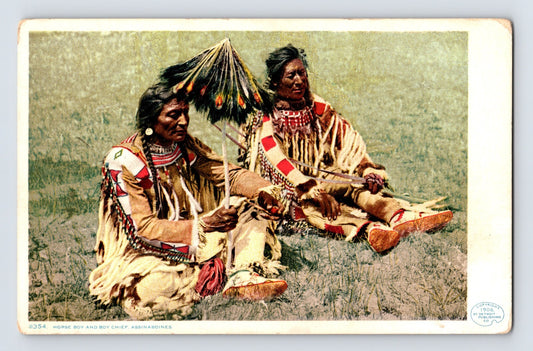 1910s colorized postcard showing two Assiniboine chiefs seated on grass in traditional regalia with beadwork, fringed garments, and feathered headdress, divided back format