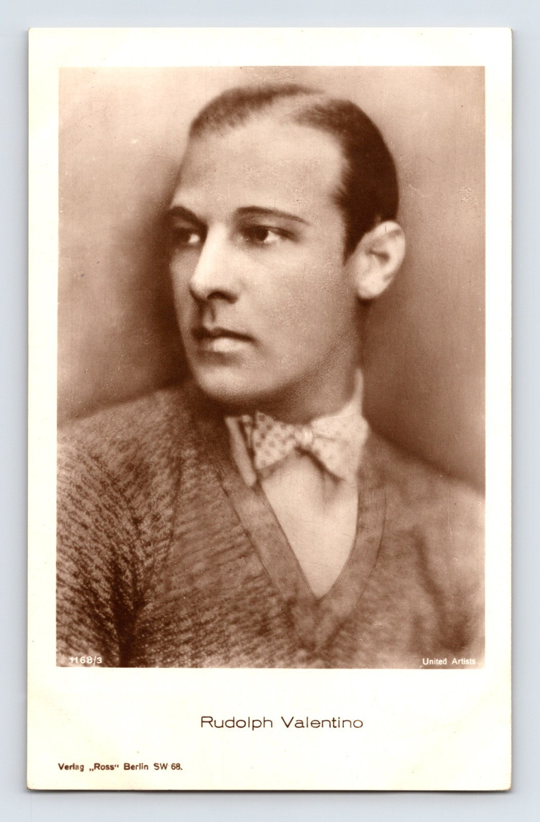 Sepia-tone portrait postcard of silent film star Rudolph Valentino wearing a patterned bow tie and knit sweater in a formal studio photograph.