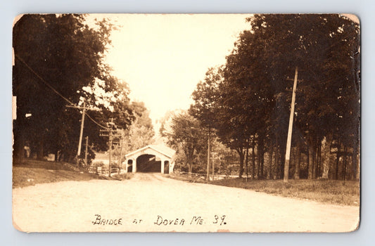 Postcard front showing a covered wooden bridge along a rural road lined with trees and telegraph poles at Dover, Maine; divided back with AZO stamp box, unposted, 1910s.