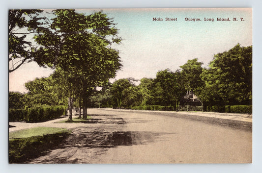 Color postcard view of Main Street in Quogue, Long Island, New York, showing a broad tree-lined residential roadway with hedges and shaded summer homes.