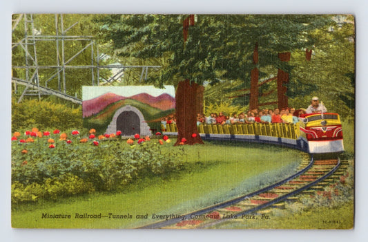 Conneaut Lake Park miniature railroad train rounding a curved track beside flowerbeds and trees, passengers seated in open cars near tunnel entrance, 1940s linen postcard Pennsylvania amusement park scene