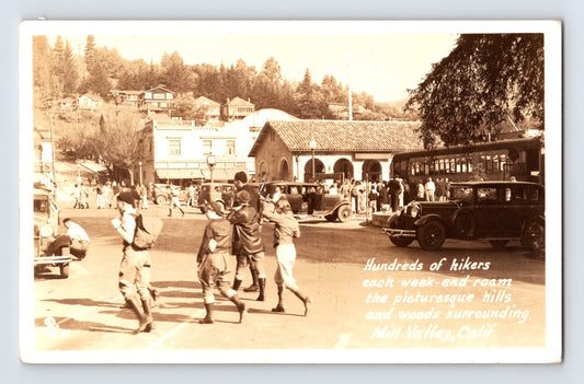 Mill Valley California 1930s RPPC postcard showing hikers crossing downtown near the Northwestern Pacific depot with early automobiles and crowds gathered along the street.