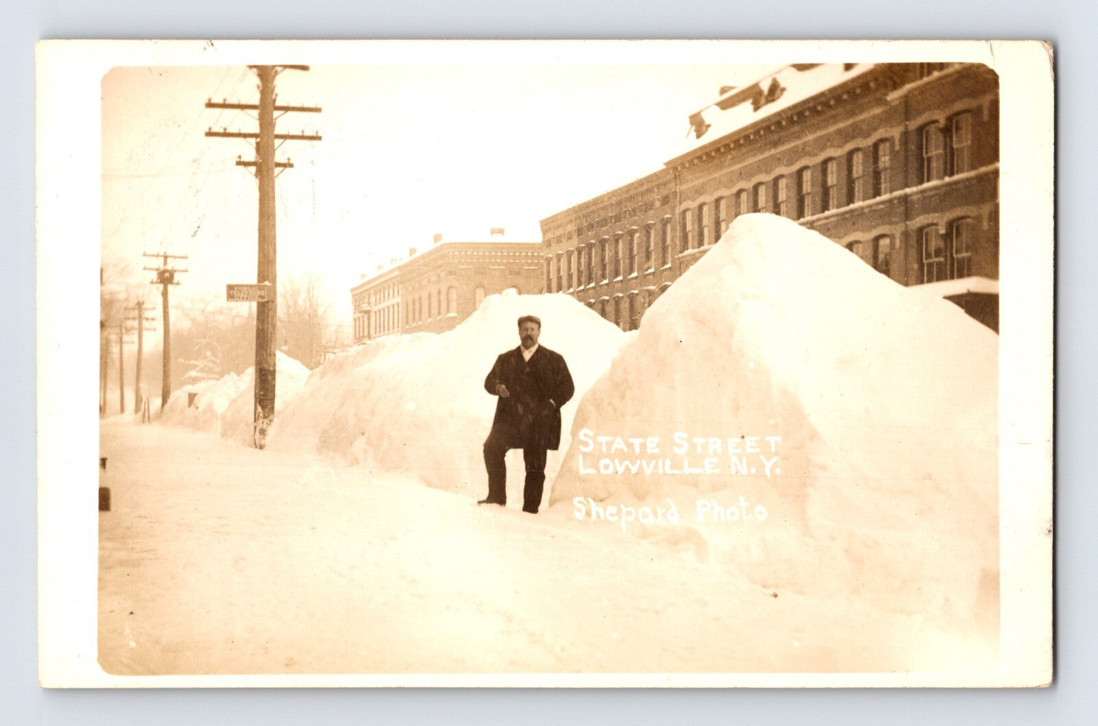 Sepia-tone winter postcard view of State Street in Lowville, New York, showing enormous snowbanks lining the street with a man standing beside the piled snow.