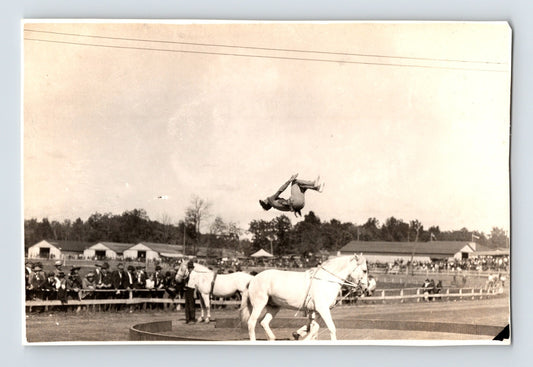 Black and white photograph showing a circus performance horse at a race track with an acrobat captured mid-air in a back flip above the animal