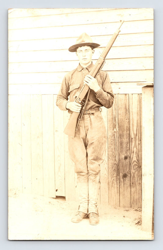 Sepia real photo postcard showing a young soldier in uniform holding a rifle, standing against a wooden building exterior; divided back with correspondence and address lines.