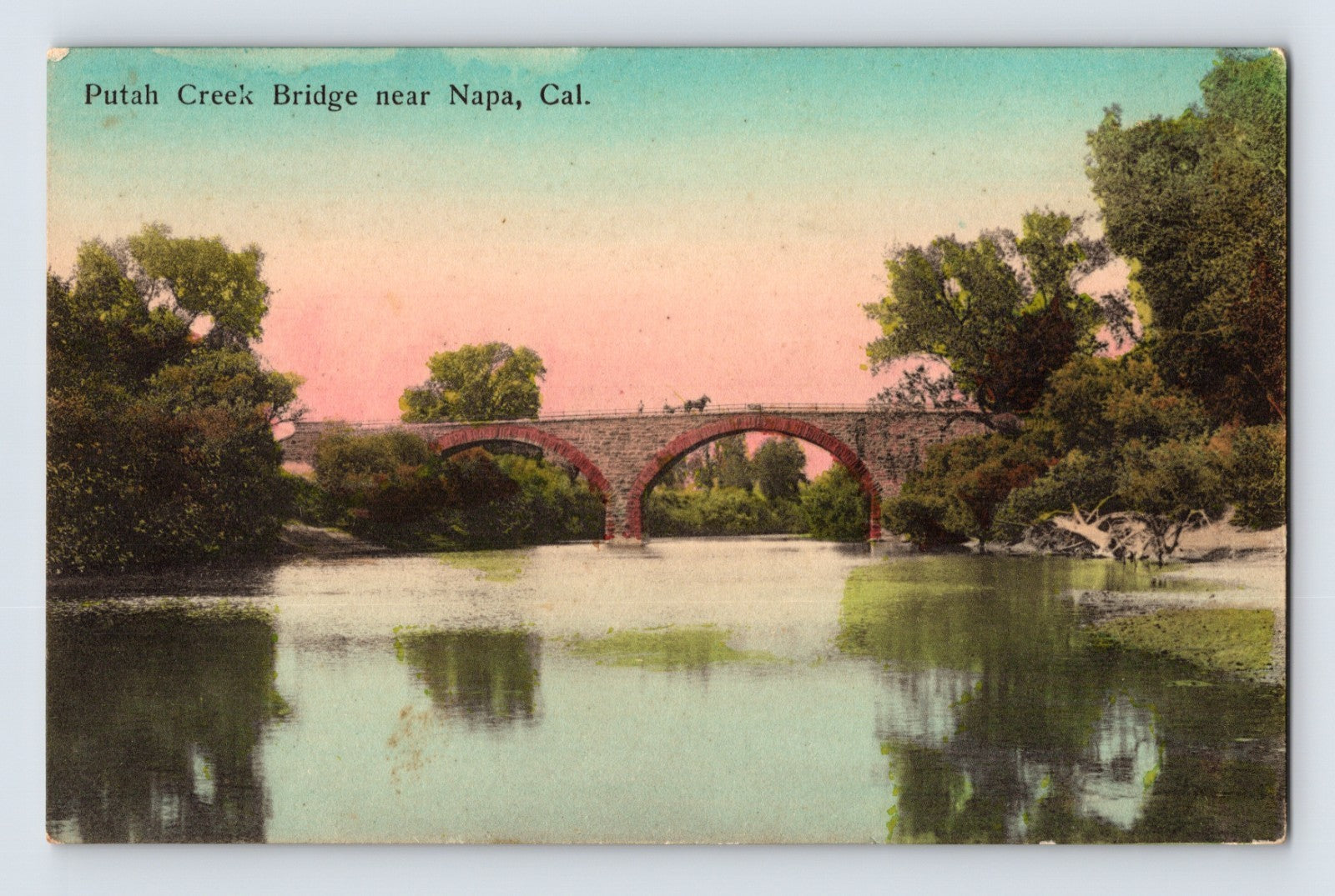 Putah Creek Bridge near Napa California postcard showing an arched masonry bridge crossing a calm waterway with reflected trees and horse-drawn traffic.