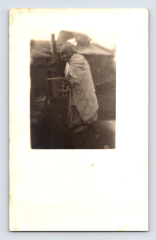 Elderly Native American woman wrapped in blanket standing beside wooden post RPPC 1910s portrait