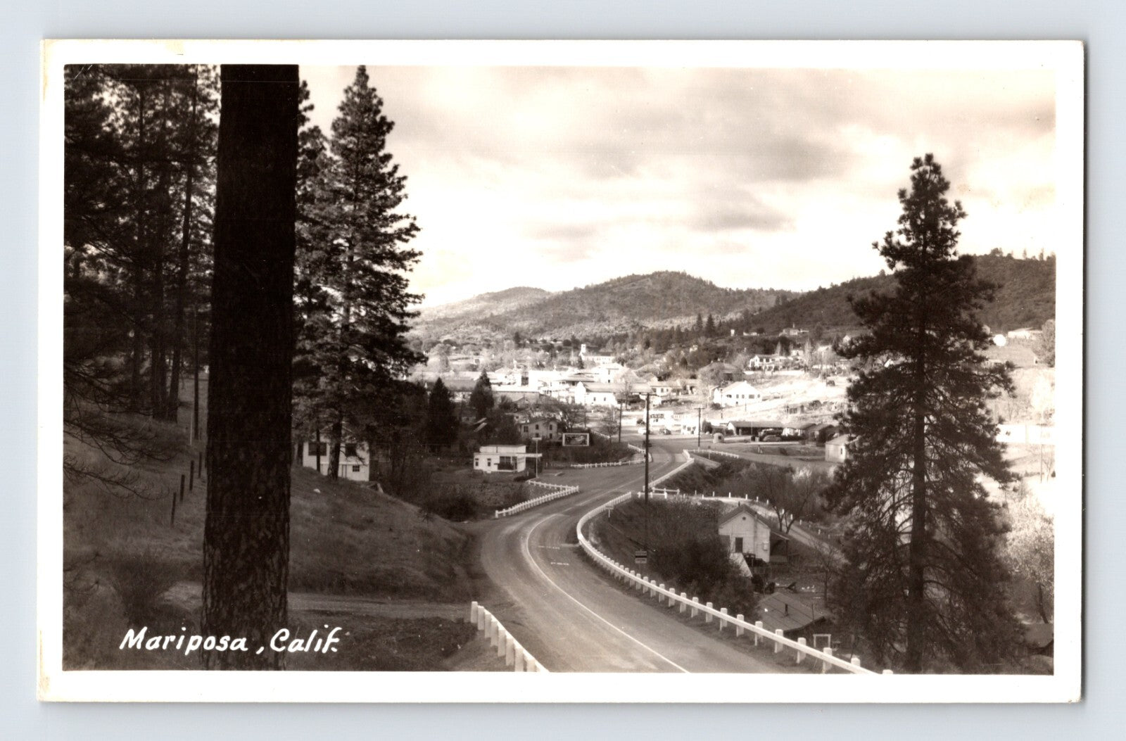 Black and white real photo postcard view of Mariposa, California with winding road leading into town, hillside homes, pine trees, and low mountains in background