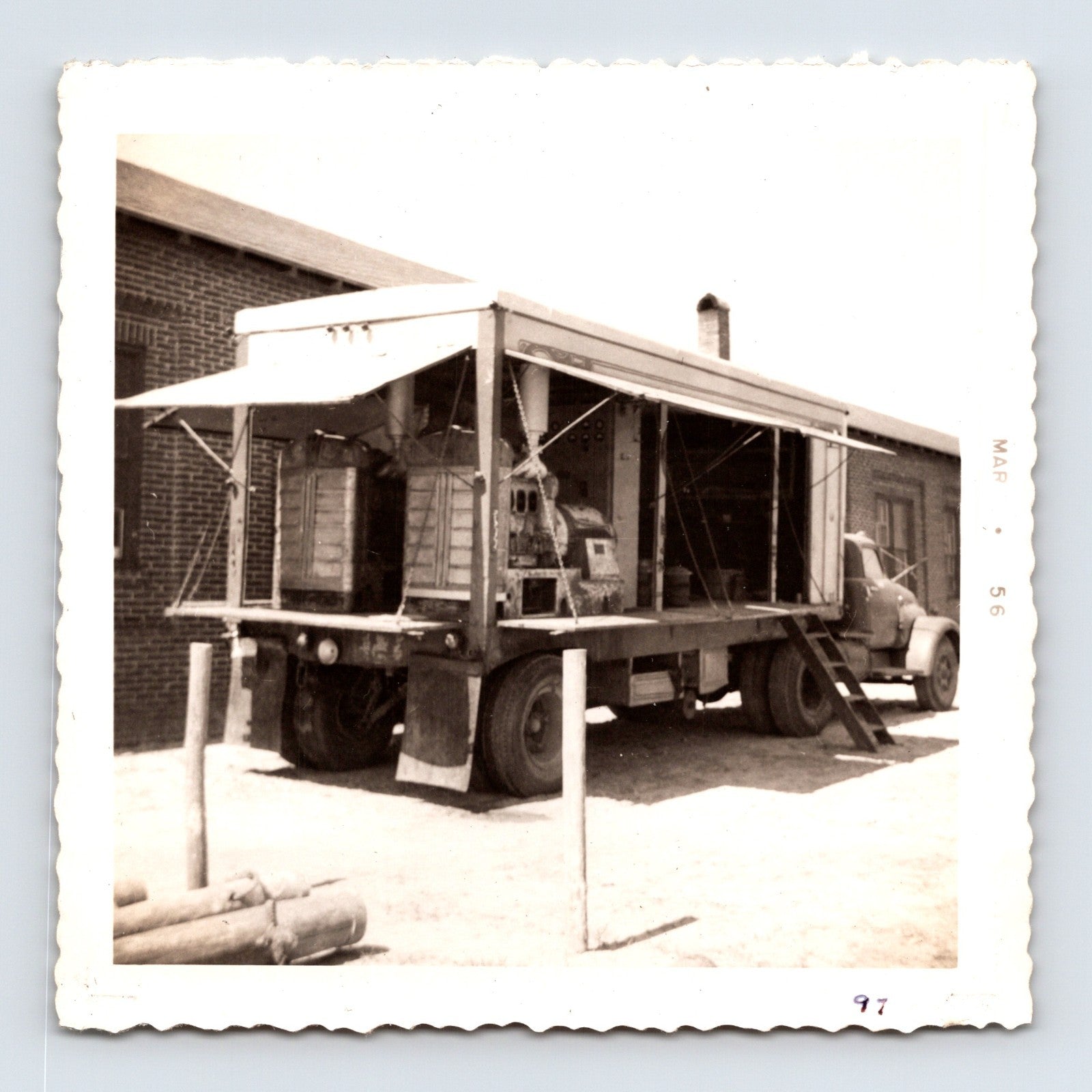 1956 circus midway equipment truck photograph showing an open-sided trailer with electrical control panels and concession machinery beside a brick service building.