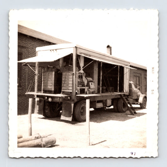 1956 circus midway equipment truck photograph showing an open-sided trailer with electrical control panels and concession machinery beside a brick service building.