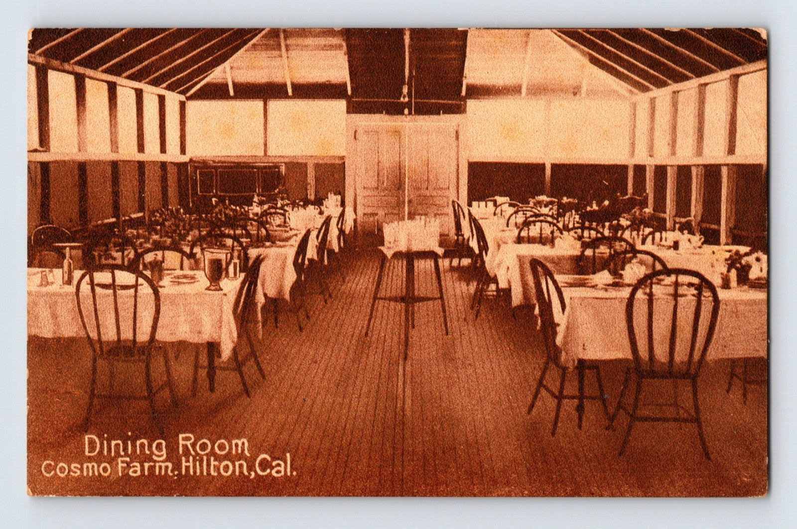 Hilton California Cosmo Farm dining room postcard showing long rustic dining hall interior with white tablecloth tables and wooden chairs arranged beneath exposed rafters.