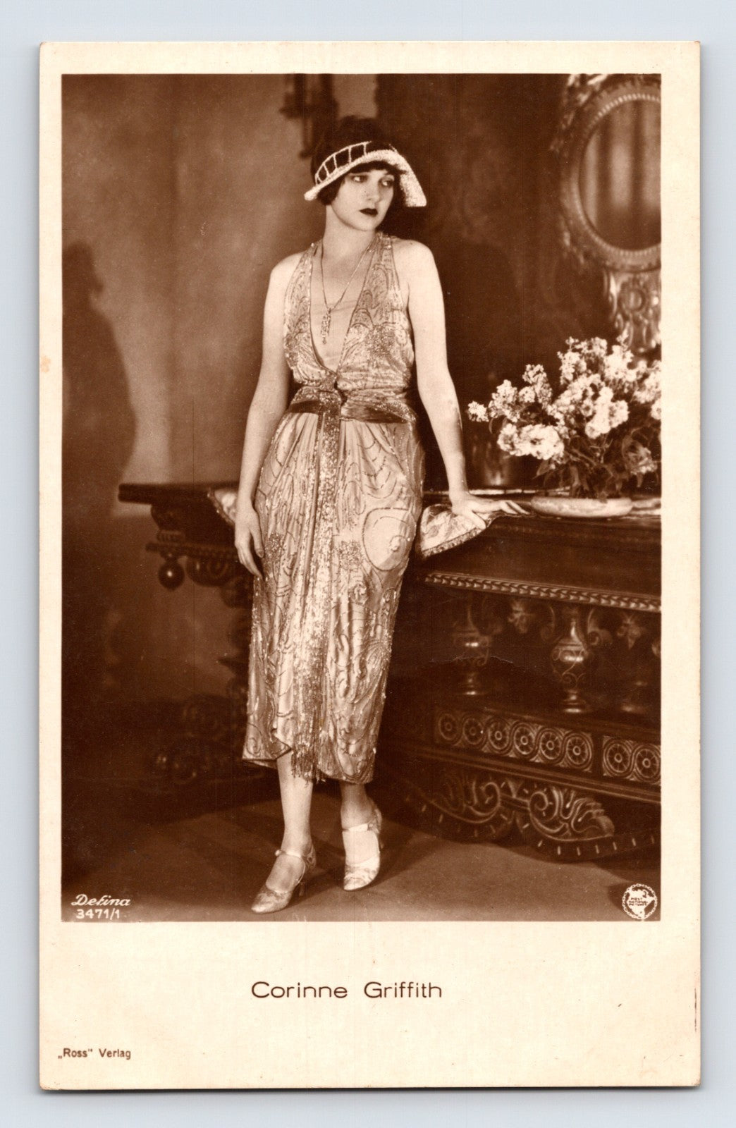 Sepia-toned real photo postcard portrait of silent film actress Corinne Griffith standing in an elegant flapper-era gown beside an ornate interior table arrangement.