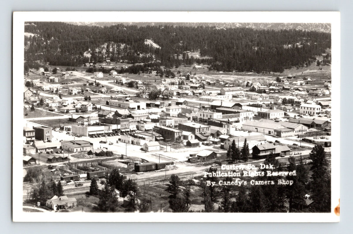 Real photo postcard aerial view of downtown Custer South Dakota with Black Hills forested slopes and Canedy Camera Shop credit on image