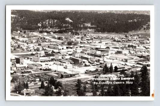 Real photo postcard aerial view of downtown Custer South Dakota with Black Hills forested slopes and Canedy Camera Shop credit on image