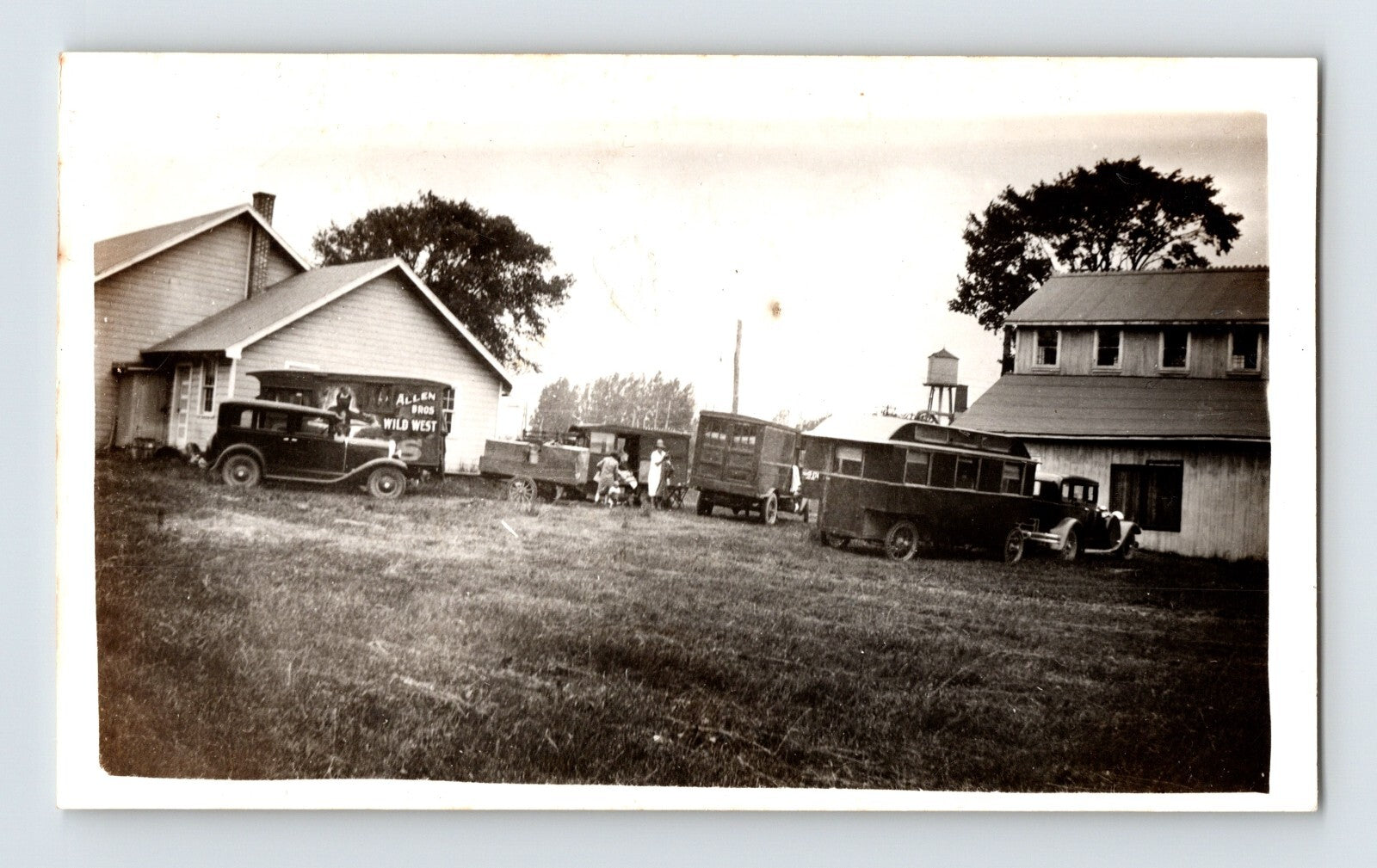 Sepia photograph of Allen Bros Wild West Show trucks parked beside rural buildings with early automobiles and equipment in Nassau New York