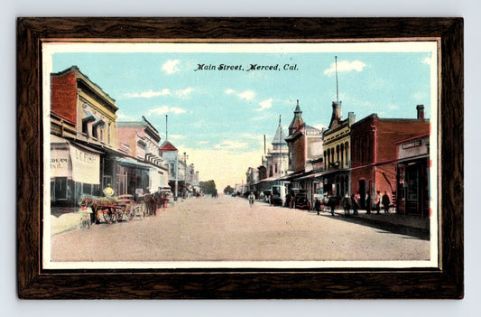1911 postcard view of Main Street in downtown Merced, California, showing early commercial buildings, horse-drawn wagons, storefronts, and pedestrians along a wide unpaved street.